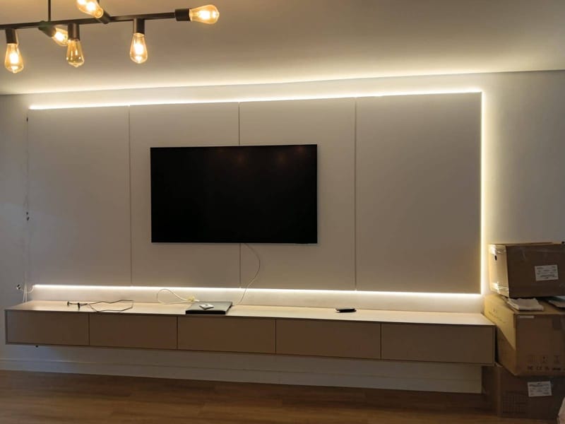 Painel de TV claro ocupando a parede inteira, com iluminação embutida no contorno e rack suspenso baixo